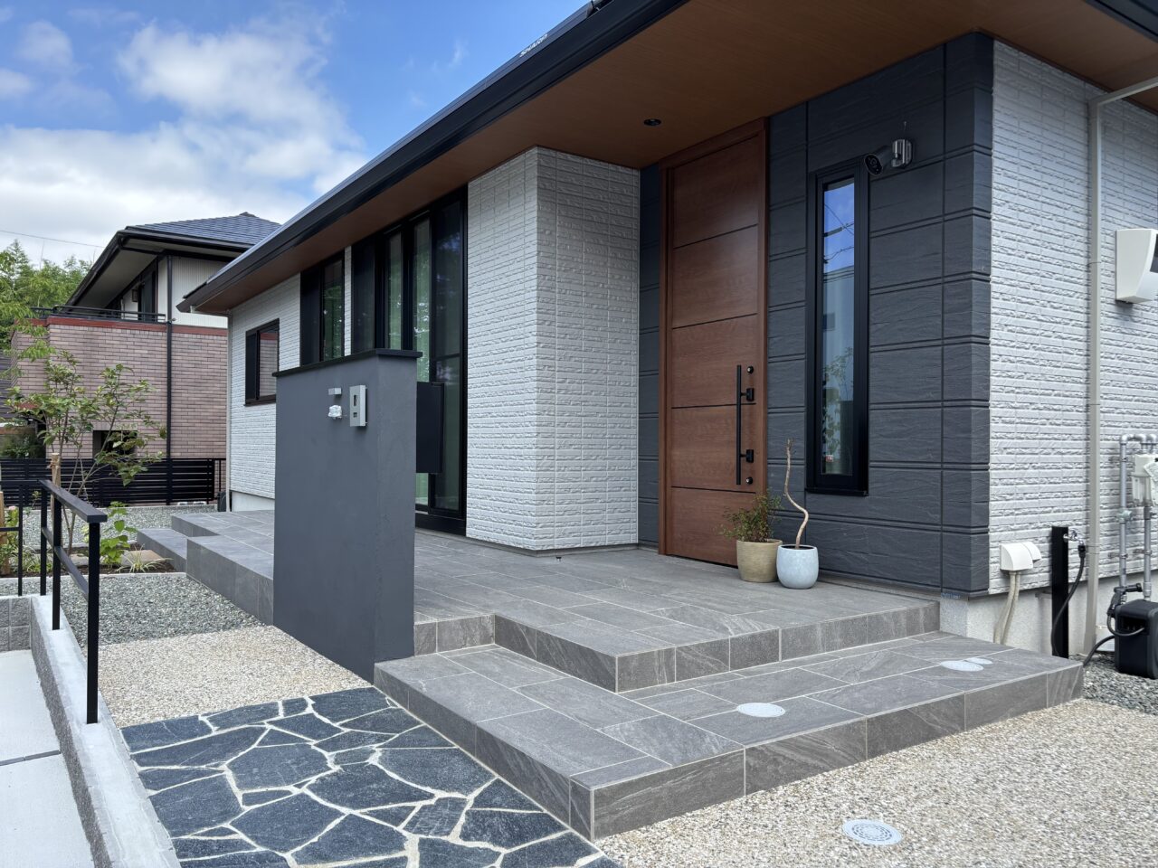 福岡市中央区の新築外構。東洋工業「オークルストーン パールブラック」で仕上げたアプローチと、落ち着いたグレーのタイルテラスが調和する玄関まわり。段差を抑えた安心設計と上質な質感が魅力の外構デザイン。｜ウェルミカ