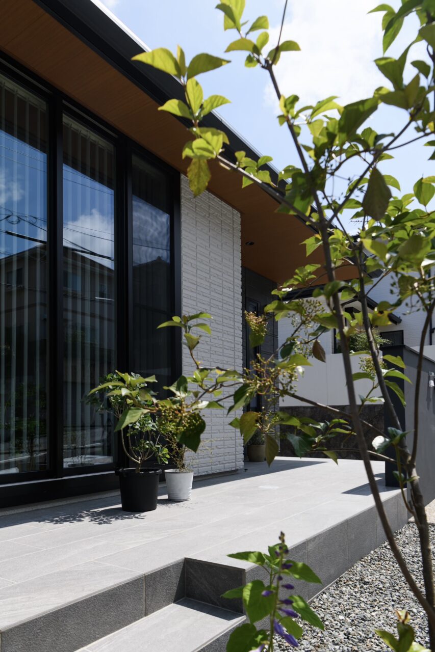 福岡市中央区の新築外構。植栽越しに見えるリビング前のタイルデッキが、平屋住宅の外観と調和し、自然を感じるやさしい印象を演出。｜ウェルミカ