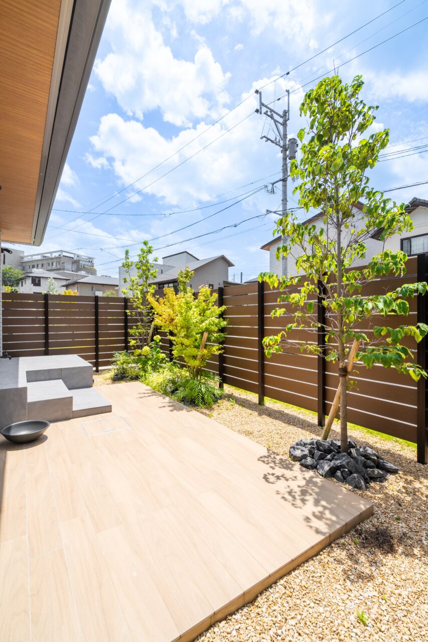 岡垣町で施工した外構リフォームの全景写真｜ブラウン系フェンスと明るいタイルデッキ、緑が調和した上質なナチュラルモダンの外構空間｜ウェルミカ