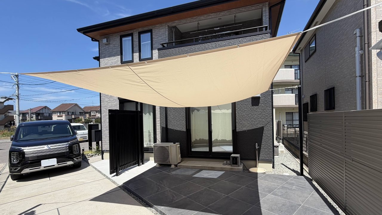 福岡市東区のお庭リフォーム事例。シックな外観の建物に調和する広々としたロータイルデッキと、日除けシェードが作り出す、上質で寛げるプライベート空間の完成写真。