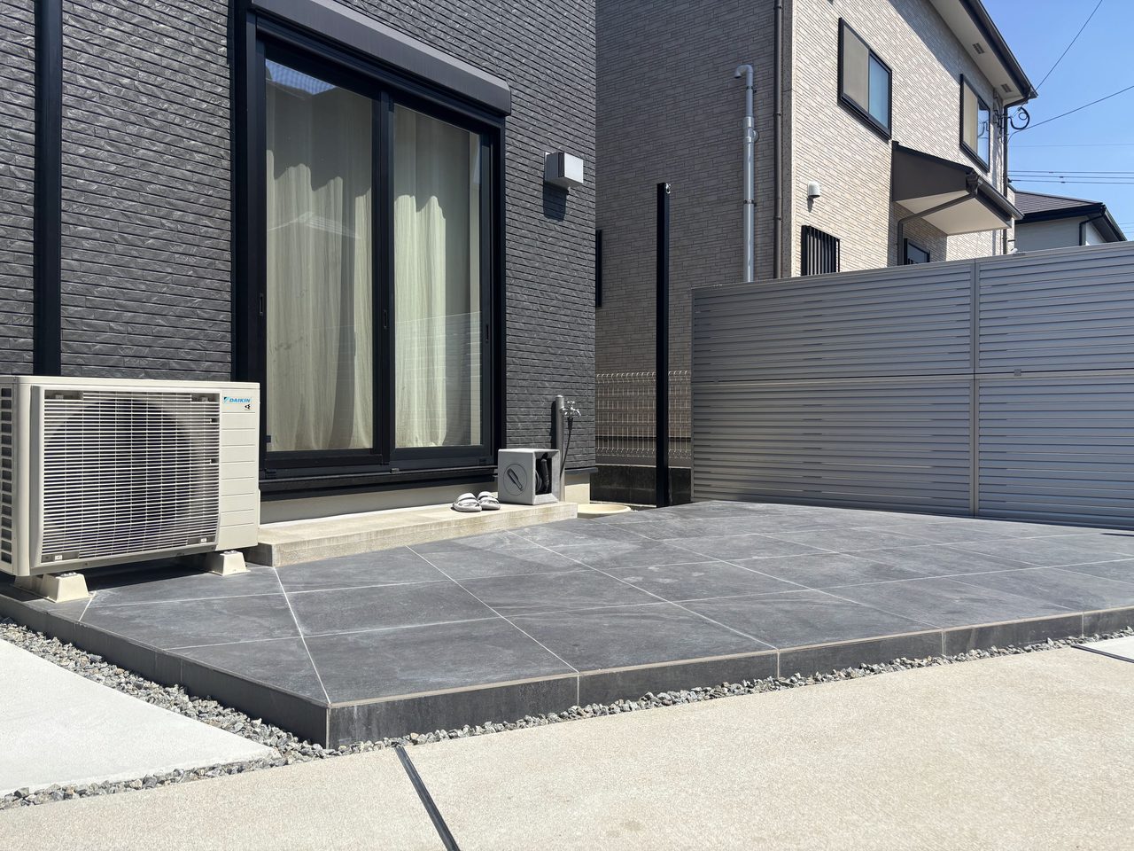 福岡市東区のお庭リフォーム施工例。お部屋から繋がりを持たせた広々とした大判のロータイルデッキと、お隣との境界に設置したシルバーの目隠しフェンス。シェード用の黒い柱も空間にすっきりと馴染んでいます。